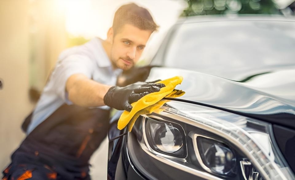 Ein Mann mit Handschuhen reinigt den Scheinwerfer eines schwarzen Autos mit einem gelben Mikrofasertuch.